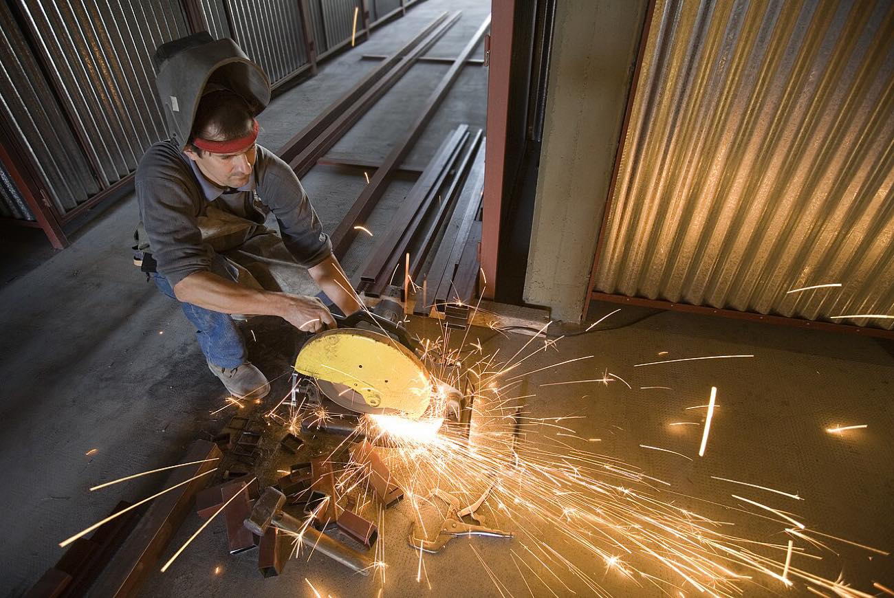 A worker crouched over a metal cut-off saw, sparks scattering across the floor, wearing no hearing protection